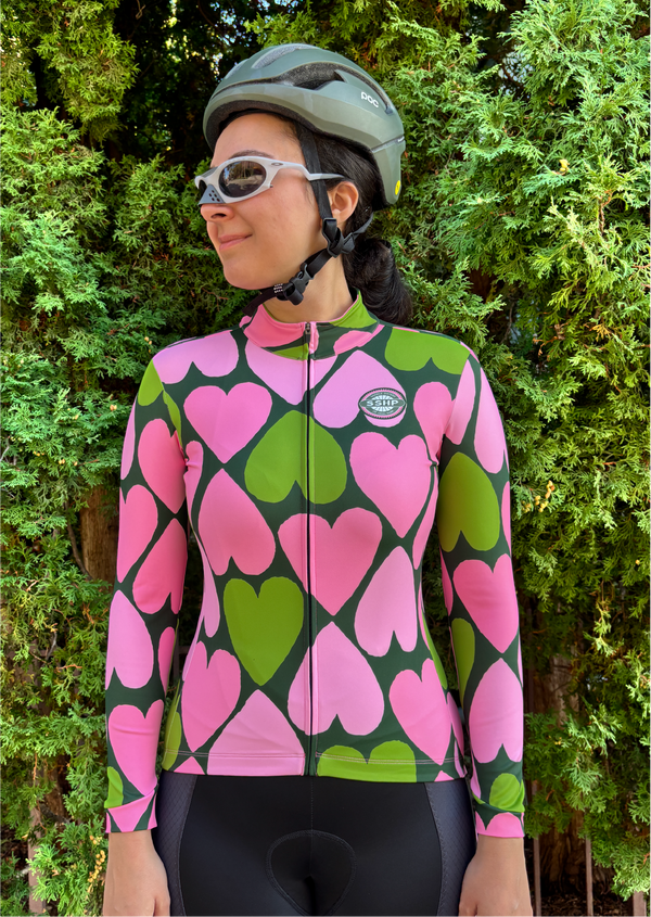 Person wearing a Daytripper and Saddle Sisters heart-patterned cycling jacket with a helmet and sunglasses outdoors.